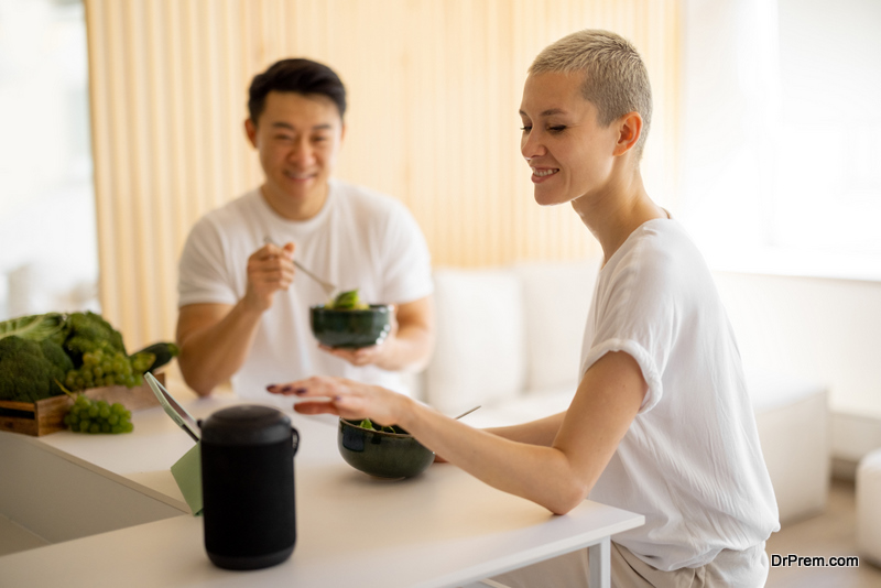 Caucasian woman pushing button on portable smart speaker while eating salad with her husband