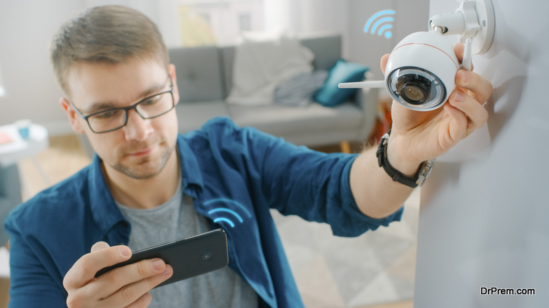 Young Man in Glasses Wearing a Blue Shirt is Adjusting a Modern Wi-Fi Surveillance Camera with Two Antennas on a White Wall at Home. He's Checking the Video Feed on his Smartphone.