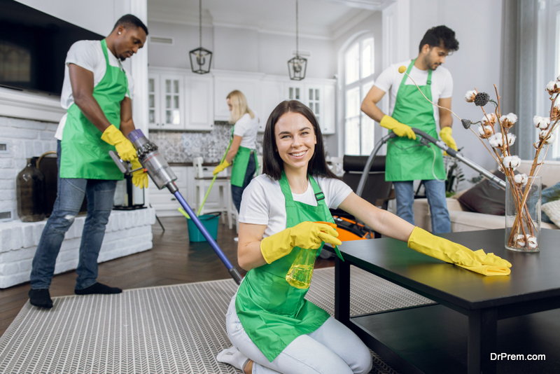 Pretty smiling Caucasian woman in green apron and yellow gloves, wiping table with microfiber cloth and detergents and smiling to camera. Cleaning team tidying up on the background.